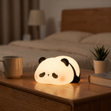 A cute panda-shaped night light glowing on a wooden bedside table in a softly lit bedroom. The lamp has a rounded white body with black ears, eyes, and cheeks, and a warm pinkish glow near the face.